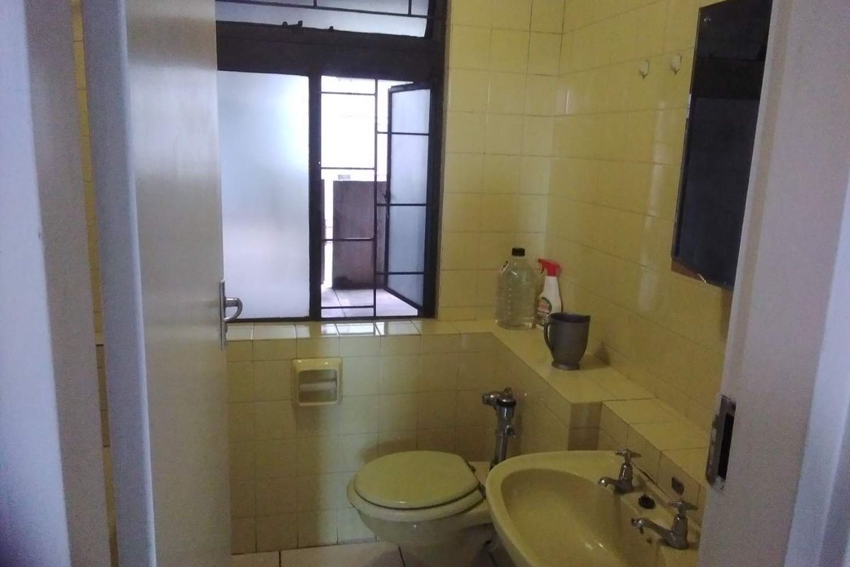 A compact bathroom featuring dated yellow wall tiles, a standard toilet, and a wall-mounted washbasin with a window providing natural light.