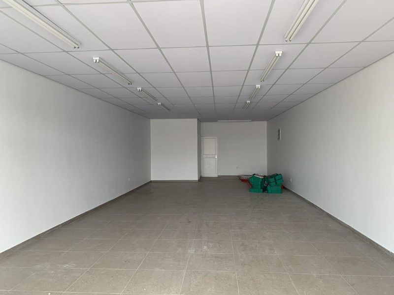 A spacious, empty retail unit featuring neutral tiled flooring, a suspended ceiling with integrated lighting, and clean white walls.