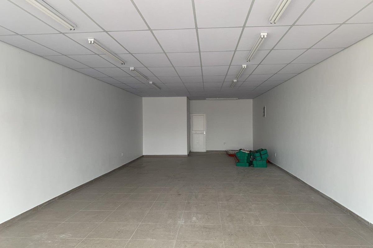 A spacious, empty retail unit featuring neutral tiled flooring, a suspended ceiling with integrated lighting, and clean white walls.
