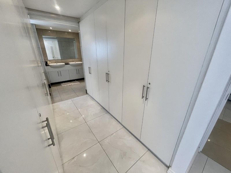 A bright hallway featuring extensive built-in storage cupboards and polished floor tiles, leading towards a modern bathroom area.