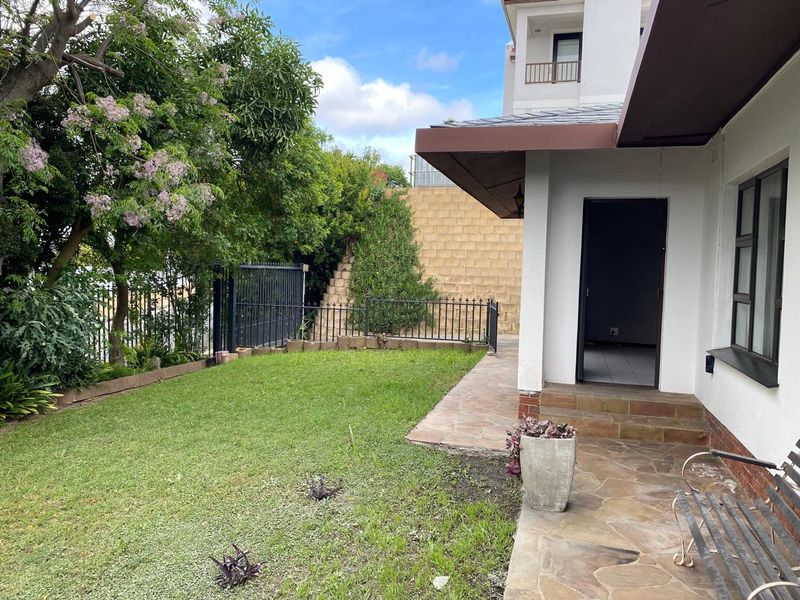 A rear exterior view of the apartment featuring a stone paved patio, a small lawn, and a retaining wall with metal perimeter fencing.