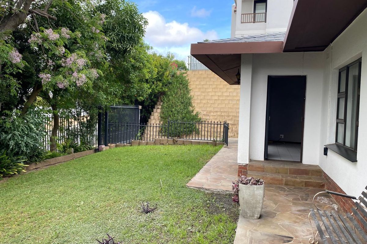 A rear exterior view of the apartment featuring a stone paved patio, a small lawn, and a retaining wall with metal perimeter fencing.