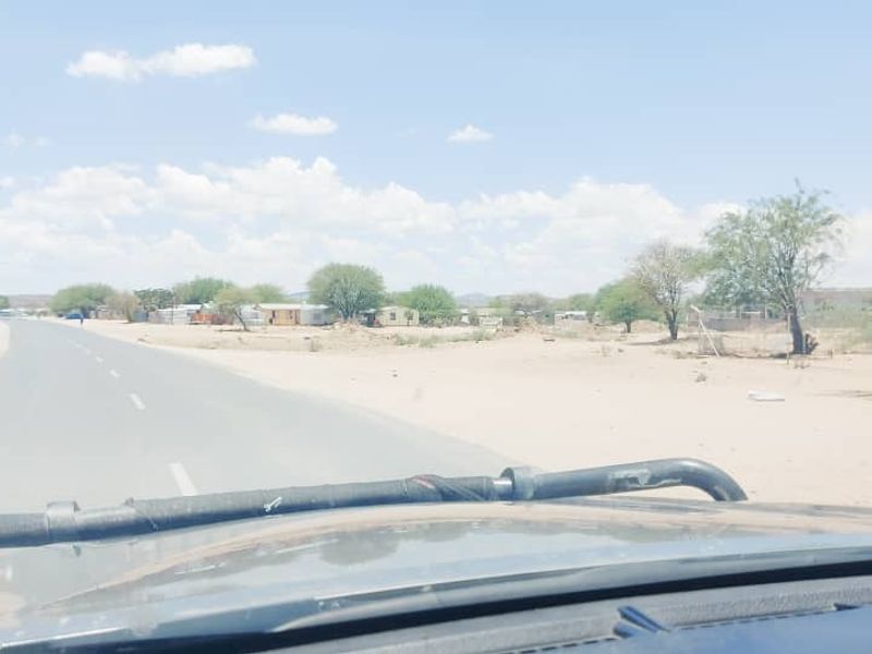 A view from a vehicle showing a paved road leading through a semi-arid landscape with sparse vegetation and distant structures.