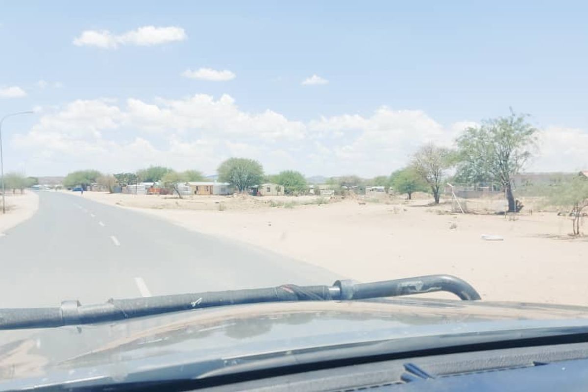A view from a vehicle showing a paved road leading through a semi-arid landscape with sparse vegetation and distant structures.