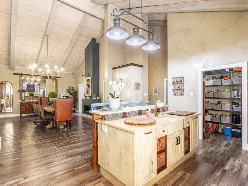 Spacious open-plan living and kitchen area featuring exposed timber beams, a central kitchen island with farmhouse sink, fireplace, and an integrated pantry.