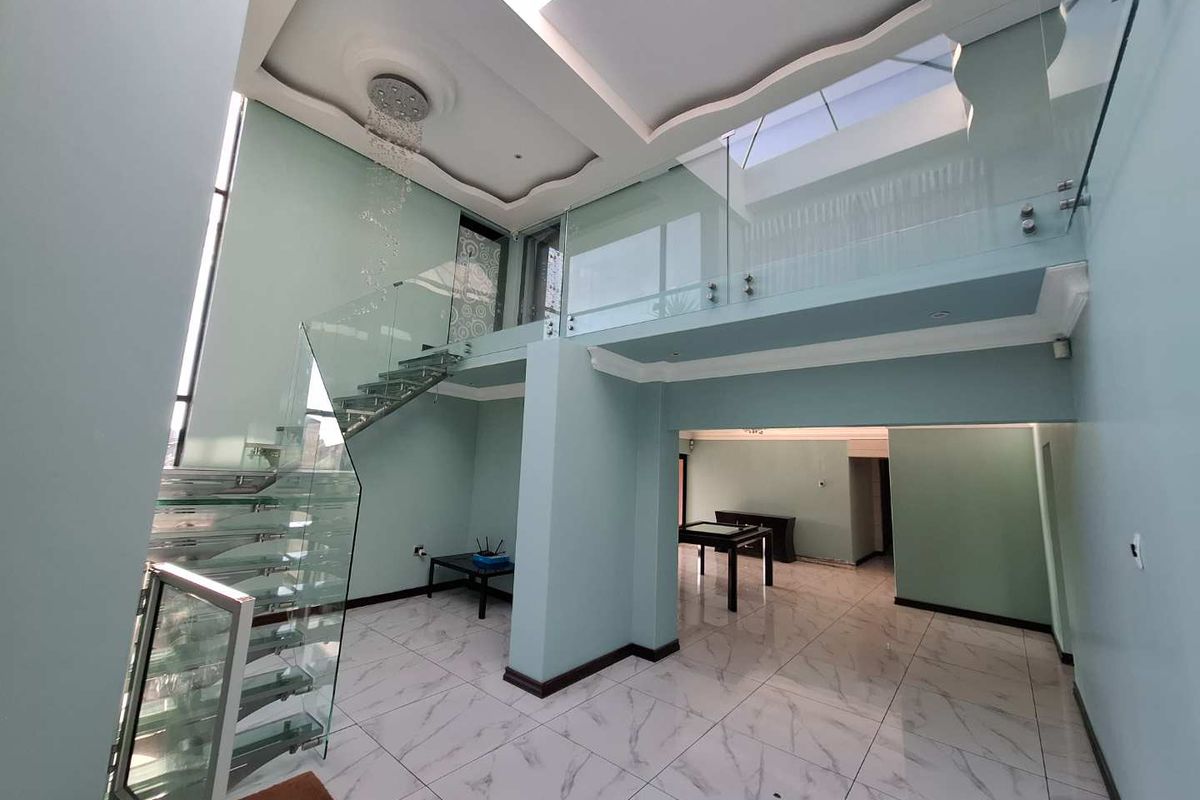 Spacious double-volume living area featuring a modern glass staircase, marble-look tiled flooring, and a mezzanine level with glass balustrades.