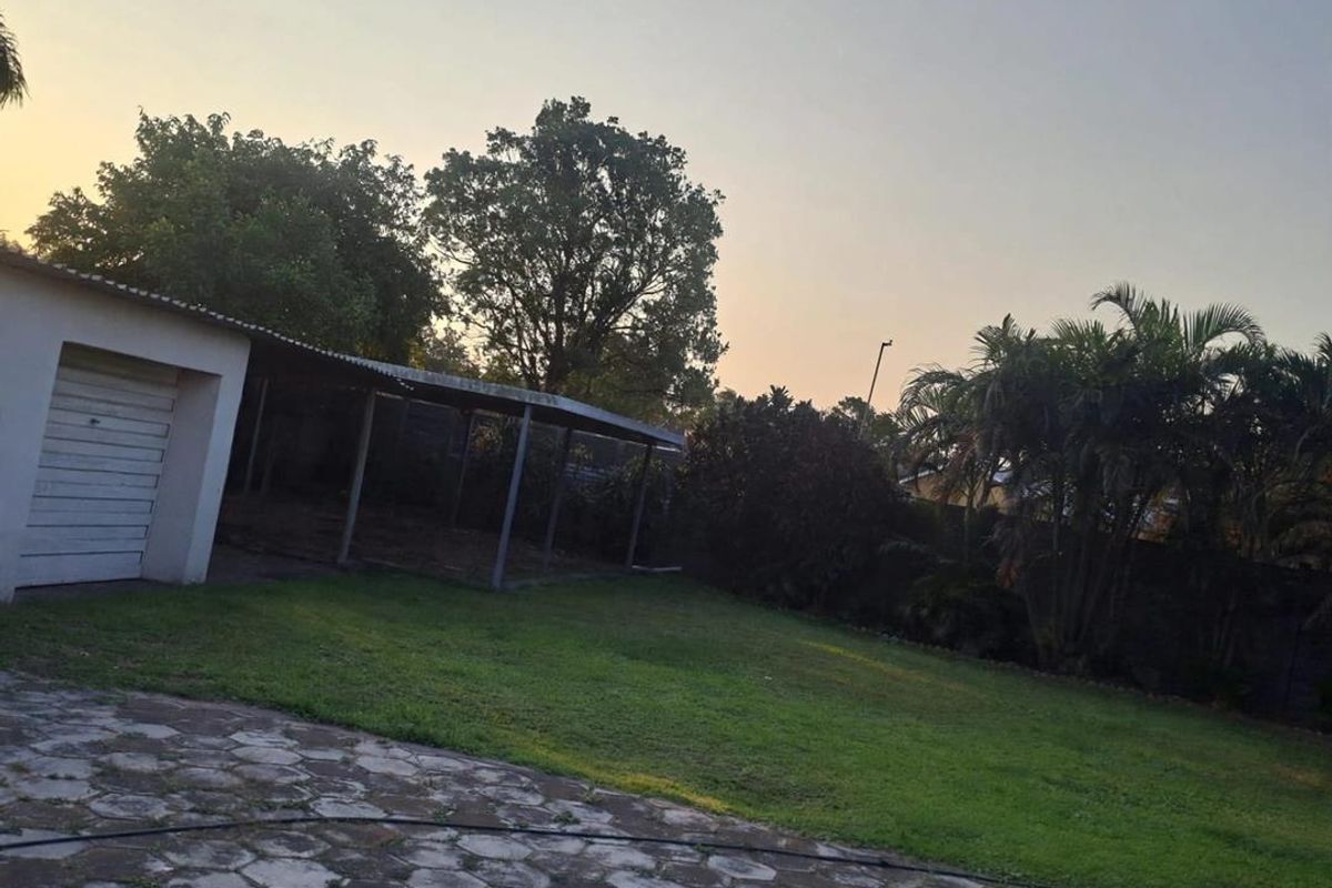 A spacious rear garden featuring a paved driveway, a single garage, and an attached carport, set against a backdrop of mature trees and boundary walls.