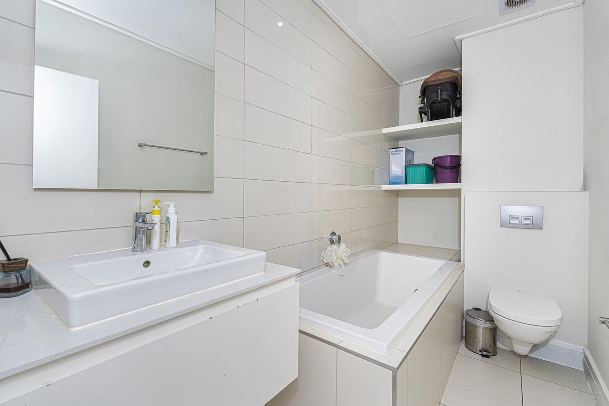 A functional bathroom featuring a built-in bathtub, wall-mounted toilet, vanity basin, and integrated shelving with neutral wall and floor tiling.