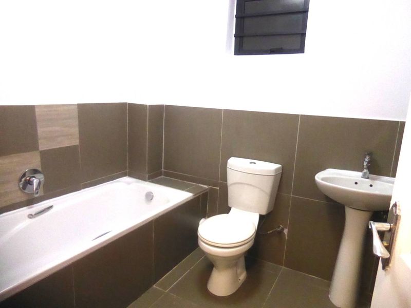 A clean, functional bathroom featuring a built-in bathtub, pedestal basin, and neutral wall tiling with a louver window for ventilation.
