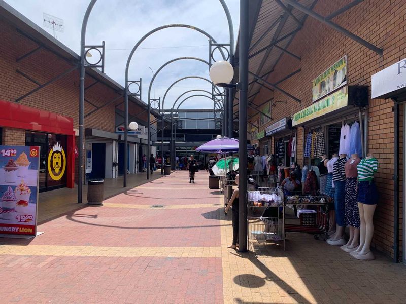 An outdoor pedestrian walkway within a retail shopping center featuring brick paving, metal archways, and various shopfronts.