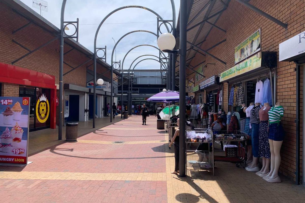 An outdoor pedestrian walkway within a retail shopping center featuring brick paving, metal archways, and various shopfronts.