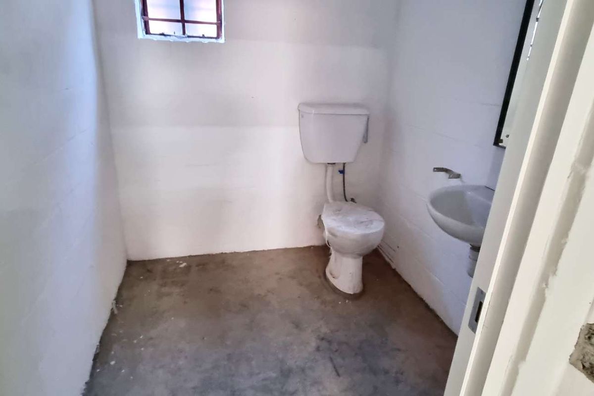 A basic industrial-style bathroom featuring a toilet, wall-mounted basin, and security-barred window, requiring significant cosmetic updates and finishing.