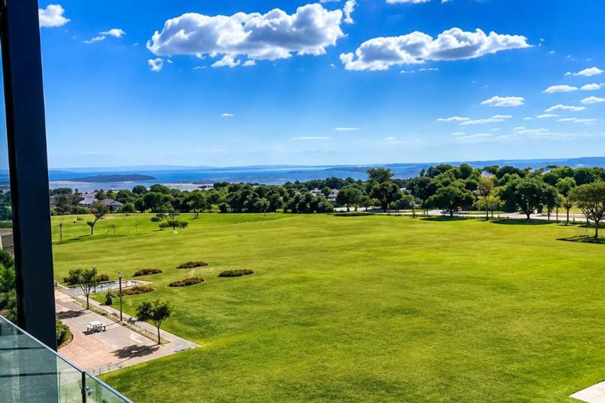 Elevated view overlooking a large green parkland area with distant water views, framed by a modern glass balcony balustrade.