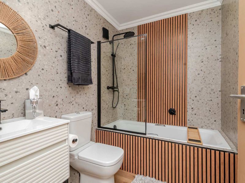 Modern bathroom featuring terrazzo-style wall tiles, a built-in bathtub with slatted timber paneling, a glass shower screen, and contemporary black fixtures.