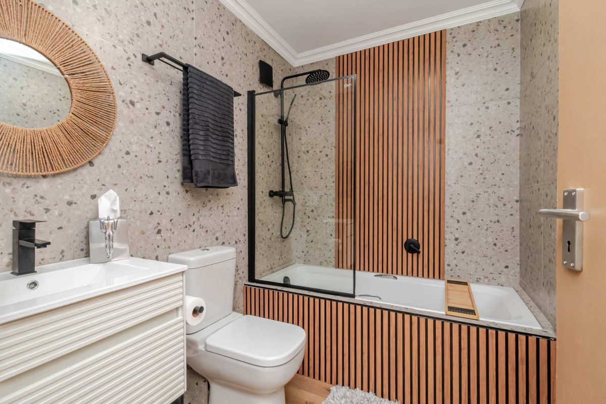 Modern bathroom featuring terrazzo-style wall tiles, a built-in bathtub with slatted timber paneling, a glass shower screen, and contemporary black fixtures.