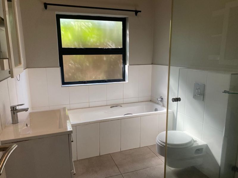 A clean, modern bathroom featuring a built-in bathtub, wall-mounted toilet, vanity basin, and a glass shower screen with neutral tiling.