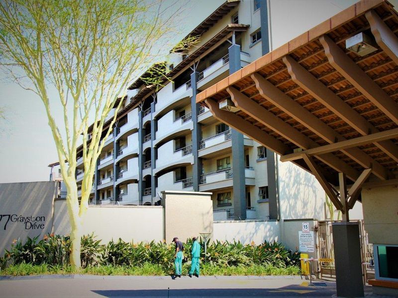 Front exterior view of the apartment complex featuring a secure entrance gate, guard checkpoint, and multi-story building facade with balconies.