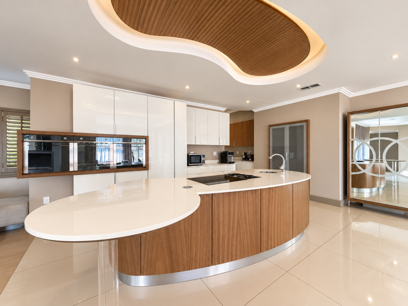 Modern kitchen featuring a large curved island with quartz countertops, high-gloss cabinetry, built-in appliances, and a unique recessed ceiling design.