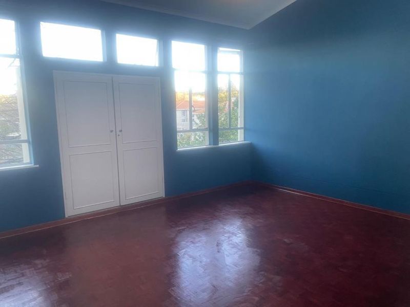 An empty bedroom featuring dark parquet flooring, blue walls, and a built-in cupboard with natural light from large windows.