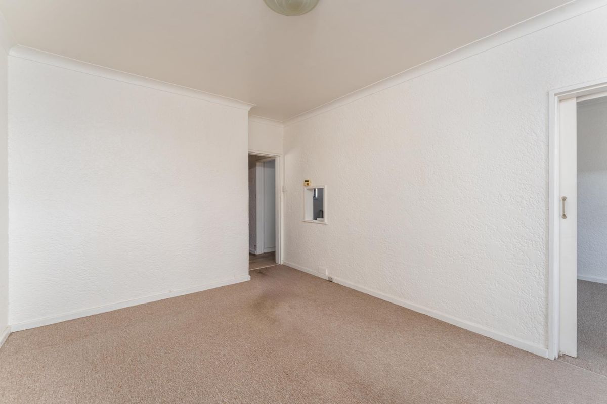 An empty, neutrally painted room featuring textured walls, beige carpeting, and a small interior service window connecting to an adjacent space.