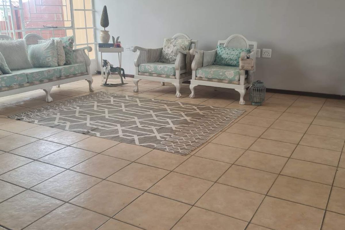 A living room area featuring neutral tiled flooring and a large window with security bars, providing natural light to the space.