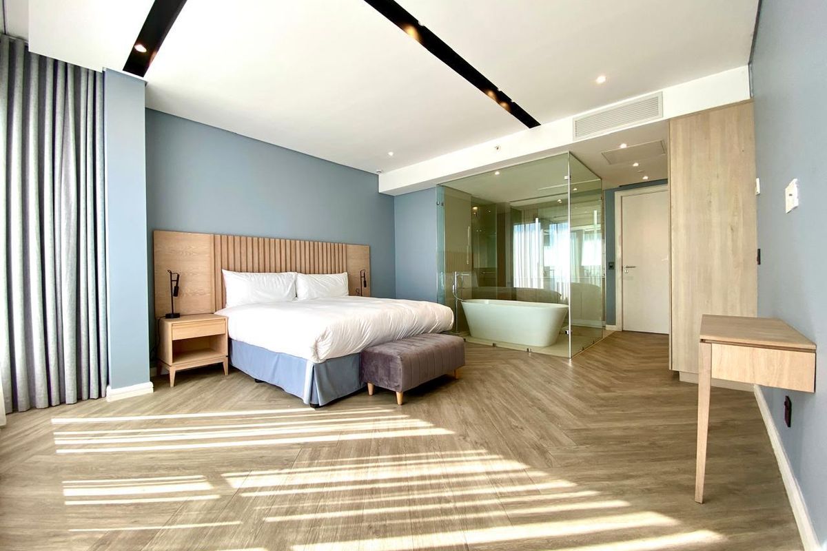 Modern bedroom featuring a glass-enclosed ensuite bathroom, herringbone wood-look flooring, built-in headboard, and recessed ceiling lighting.