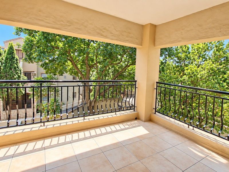 A spacious covered balcony featuring tiled flooring and a decorative wrought iron balustrade, offering a pleasant outlook over mature trees.