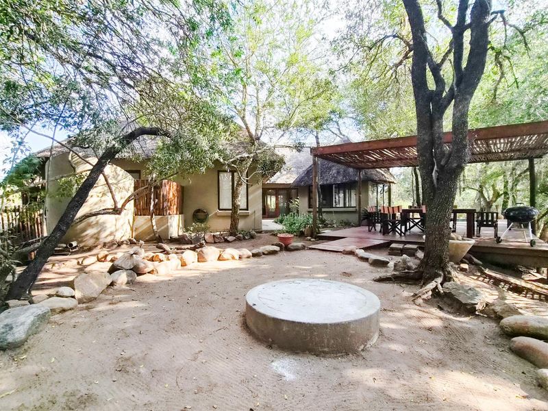Rear exterior view featuring a thatched roof dwelling, a raised timber deck with pergola, and a central concrete fire pit in a natural garden setting.
