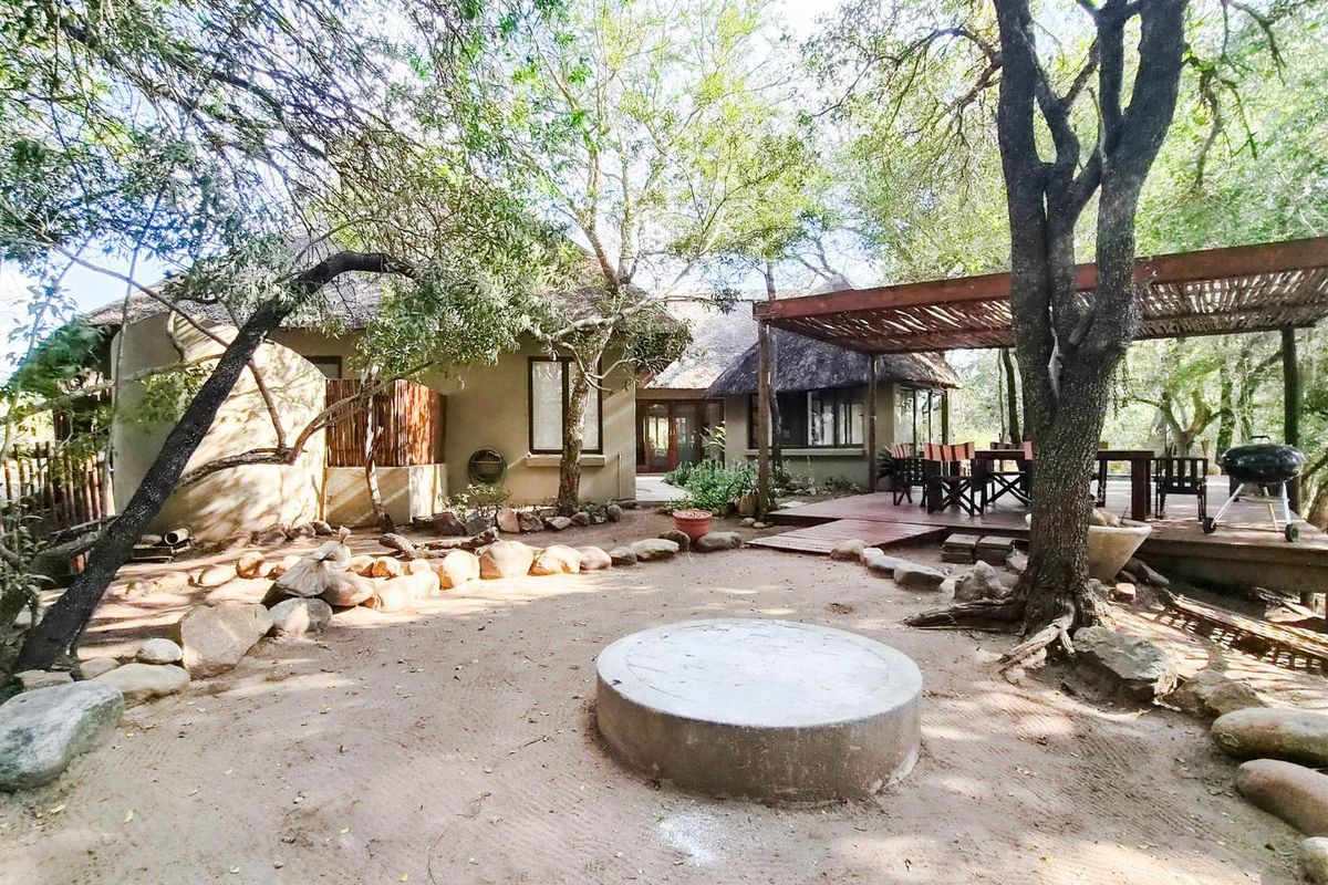 Rear exterior view featuring a thatched roof dwelling, a raised timber deck with pergola, and a central concrete fire pit in a natural garden setting.
