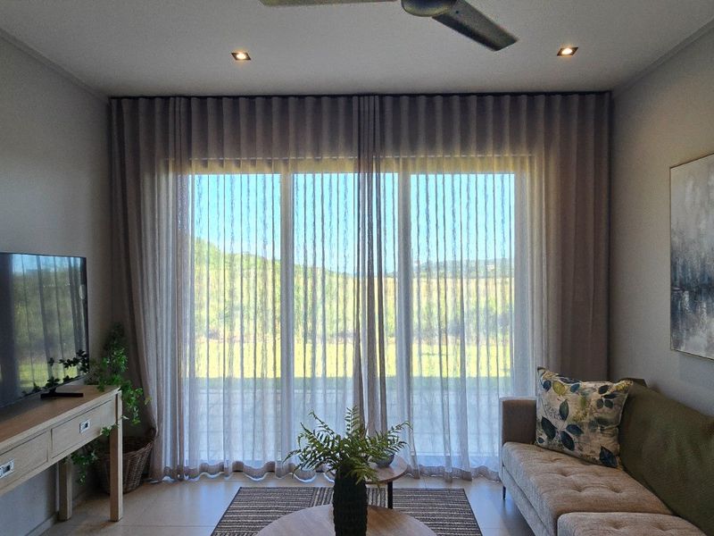 A well-maintained living room featuring tiled flooring, recessed downlighting, a ceiling fan, and large sliding glass doors with elegant sheer curtains.