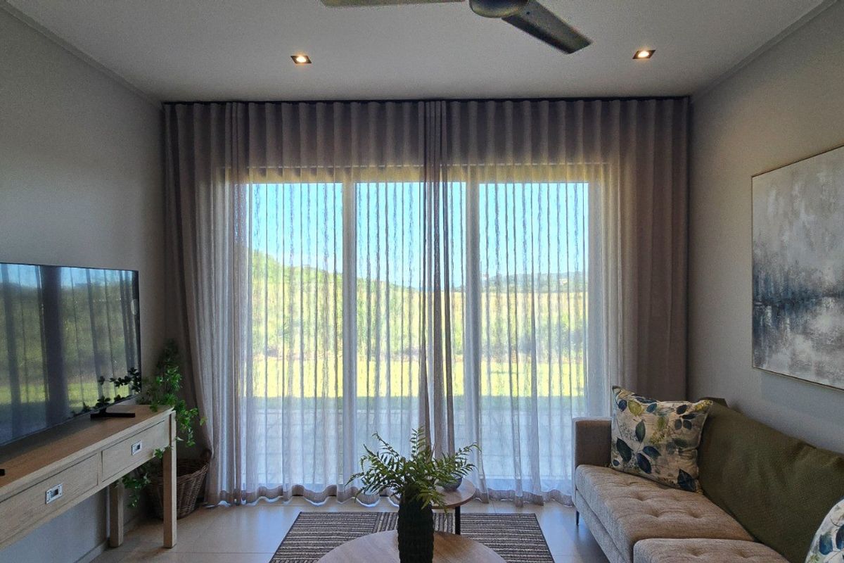 A well-maintained living room featuring tiled flooring, recessed downlighting, a ceiling fan, and large sliding glass doors with elegant sheer curtains.