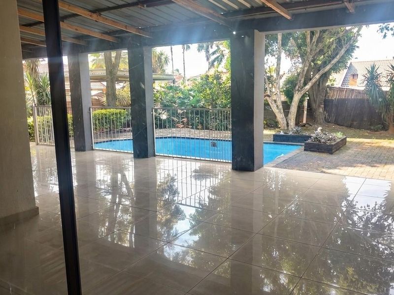 A spacious covered patio featuring polished floor tiles, exposed timber rafters, and a metal roof, overlooking a private swimming pool and garden area.