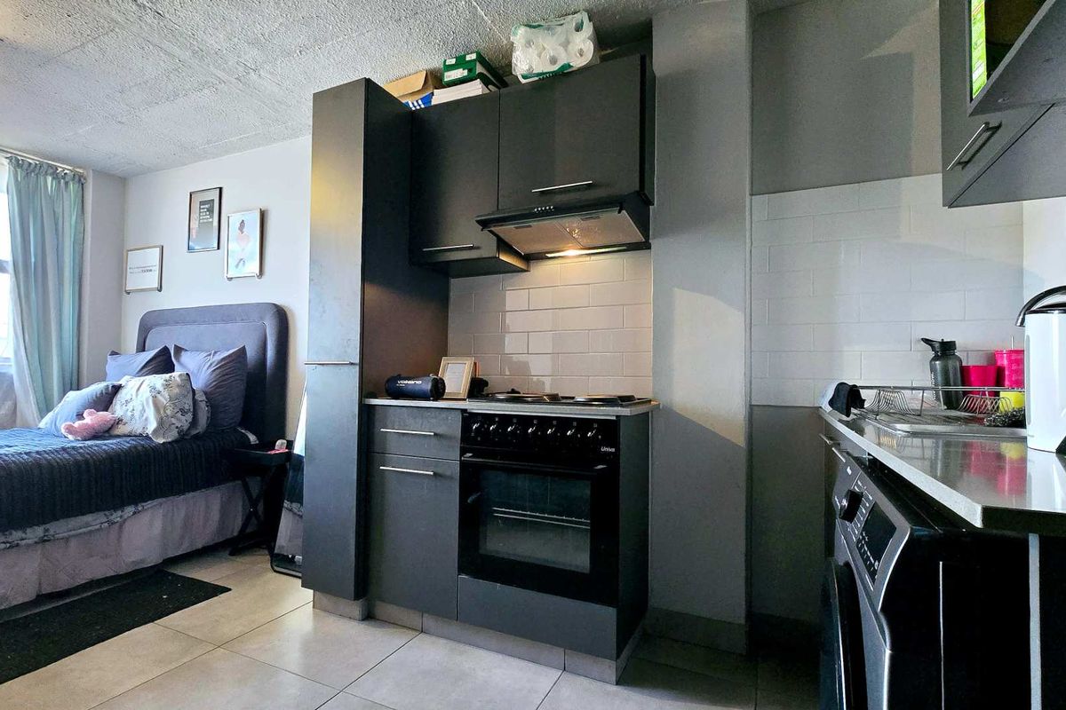 Compact open-plan studio apartment featuring a modern kitchenette with built-in oven, hob, extractor fan, and subway tile backsplash, seamlessly integrated with the sleeping area.