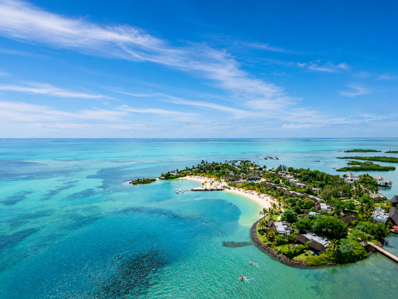 An expansive aerial view of a tropical island resort featuring lush landscaping, private villas, a sandy beach, and clear turquoise ocean waters.