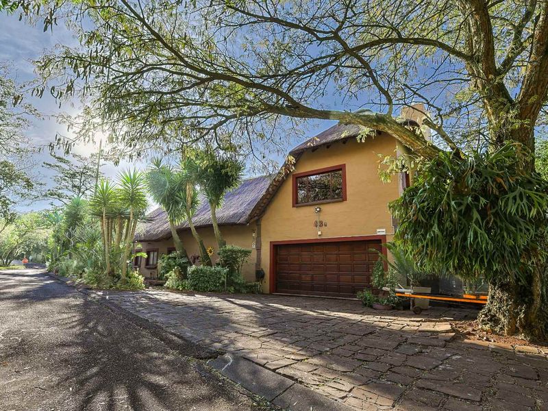 Front exterior view of a charming thatched-roof house featuring a paved driveway and integrated garage, surrounded by mature trees.