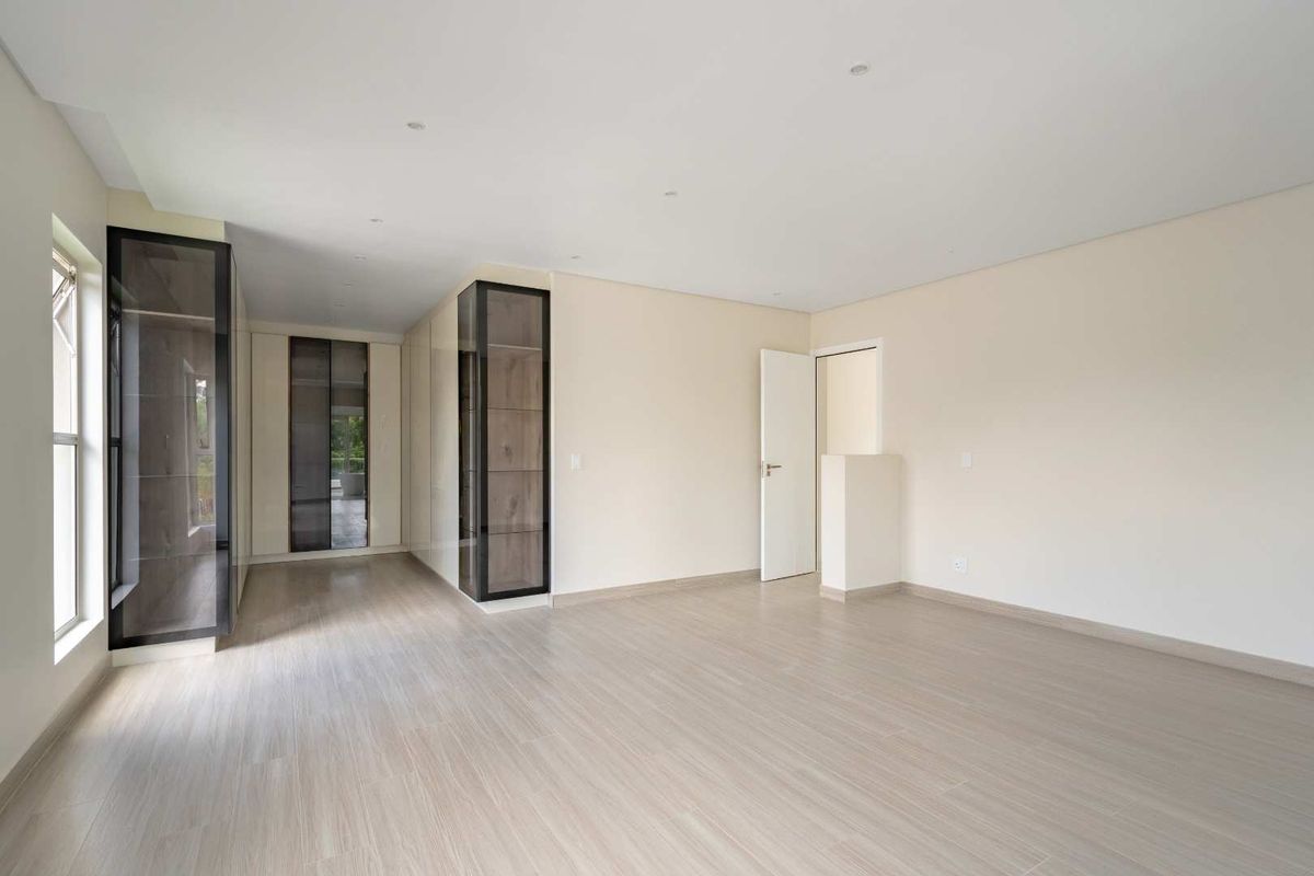 A bright, modern bedroom featuring light wood-look flooring, neutral walls, and extensive built-in cabinetry with glass-fronted display sections.