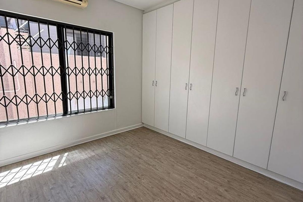 A bright, empty bedroom featuring light wood-look flooring, extensive built-in white cabinetry, a wall-mounted air conditioning unit, and security bars on the window.