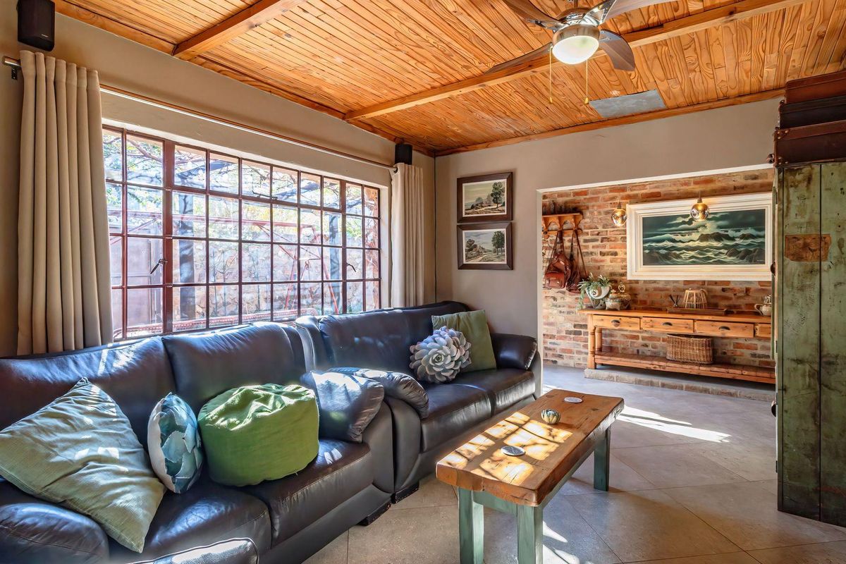 A cozy living room featuring an exposed timber ceiling, ceiling fan, tiled flooring, and a characterful exposed brick wall with large window.