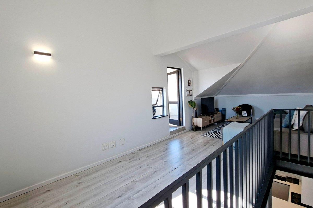 A bright living area featuring light-toned laminate flooring, a modern metal balustrade, and a unique sloped ceiling design in an apartment setting.