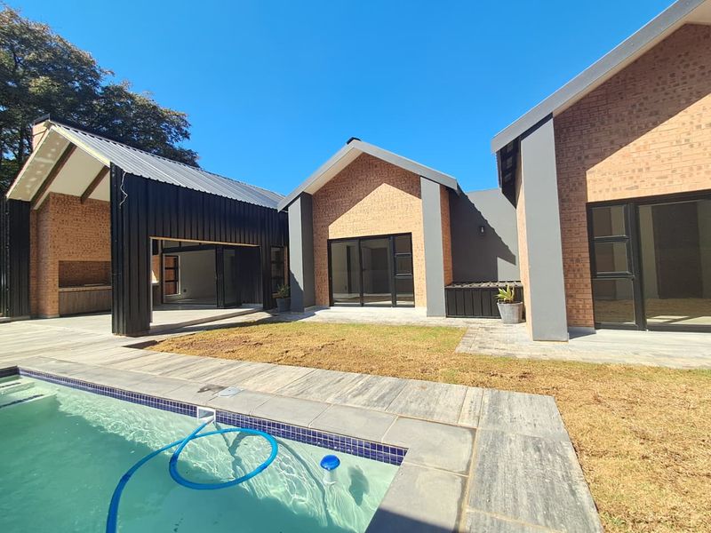 Modern rear exterior featuring a swimming pool, paved deck, and a covered patio area with a built-in braai, set against a contemporary brick facade.