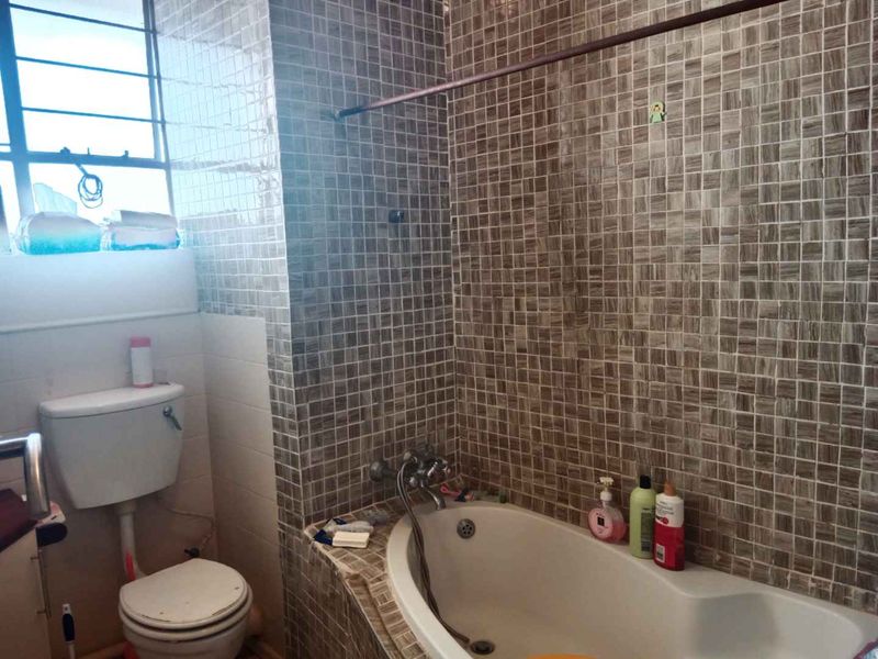 A functional bathroom featuring a built-in bathtub with mosaic wall tiling, a standard toilet, and a window for natural light.