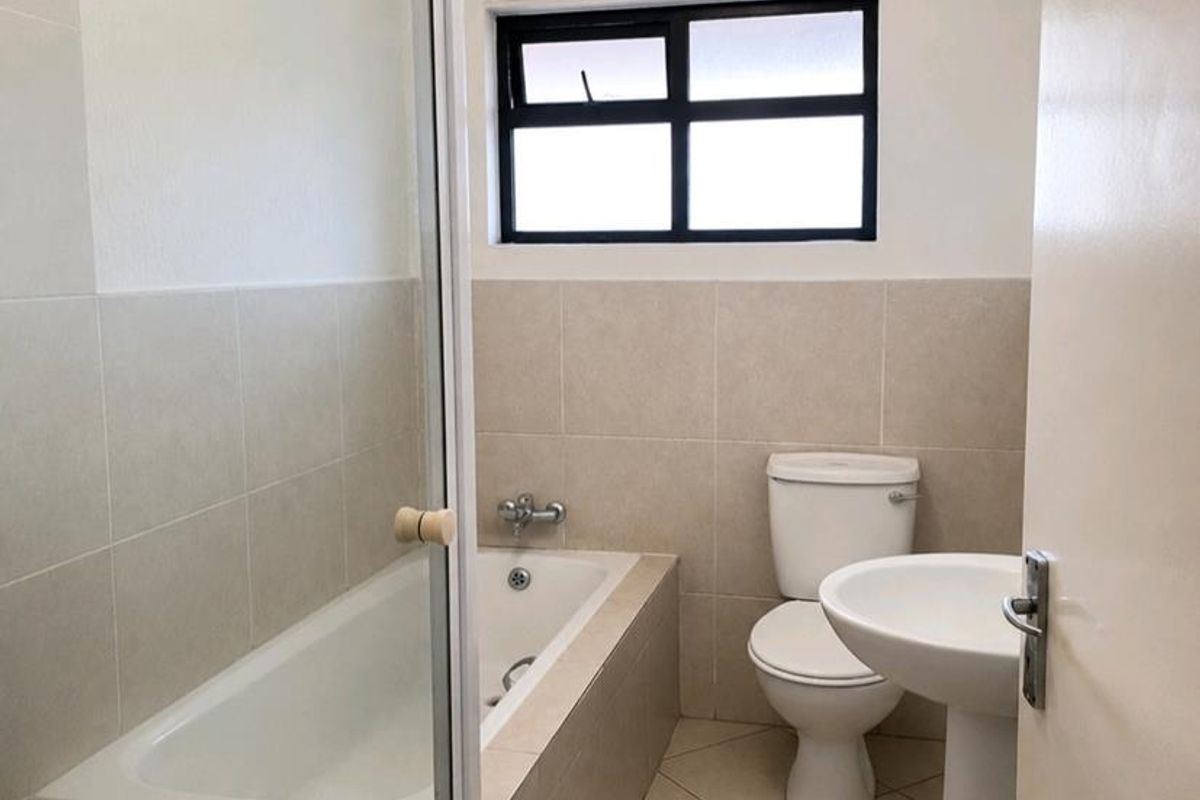 A clean, functional bathroom featuring a built-in bathtub with a glass screen, a pedestal sink, and a toilet, finished with neutral floor and wall tiles.