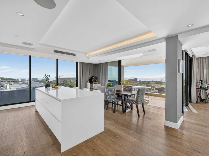 Modern open-plan living and kitchen area featuring a sleek central island, hardwood floors, and expansive floor-to-ceiling windows with seamless access to the balcony.