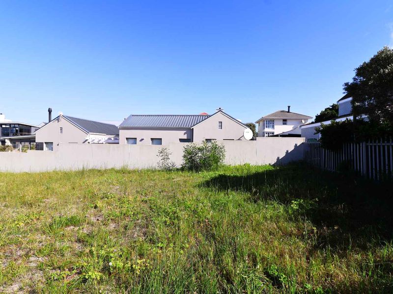 A vacant residential plot featuring natural grass coverage, enclosed by a perimeter wall and metal fencing in a suburban setting.