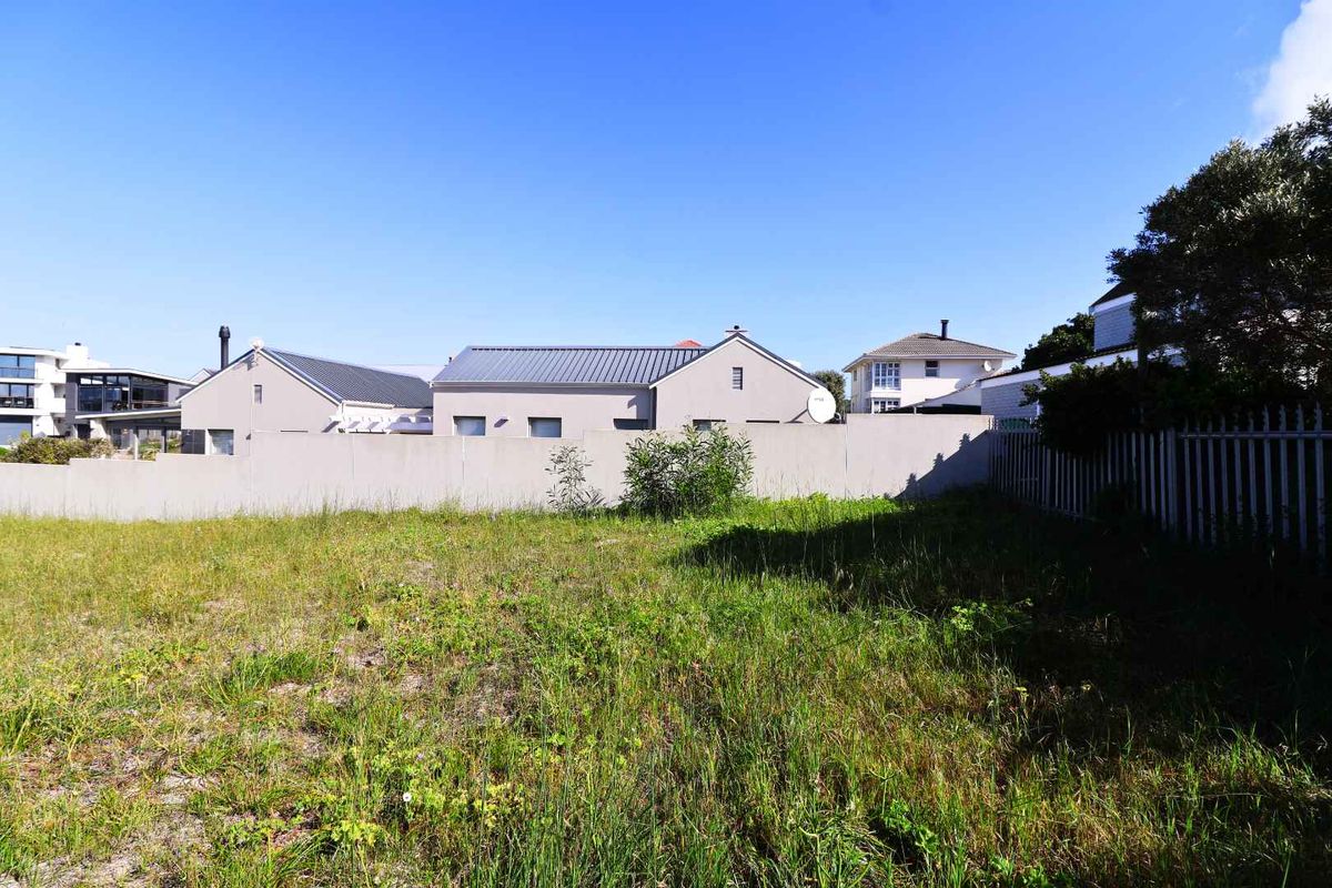 A vacant residential plot featuring natural grass coverage, enclosed by a perimeter wall and metal fencing in a suburban setting.