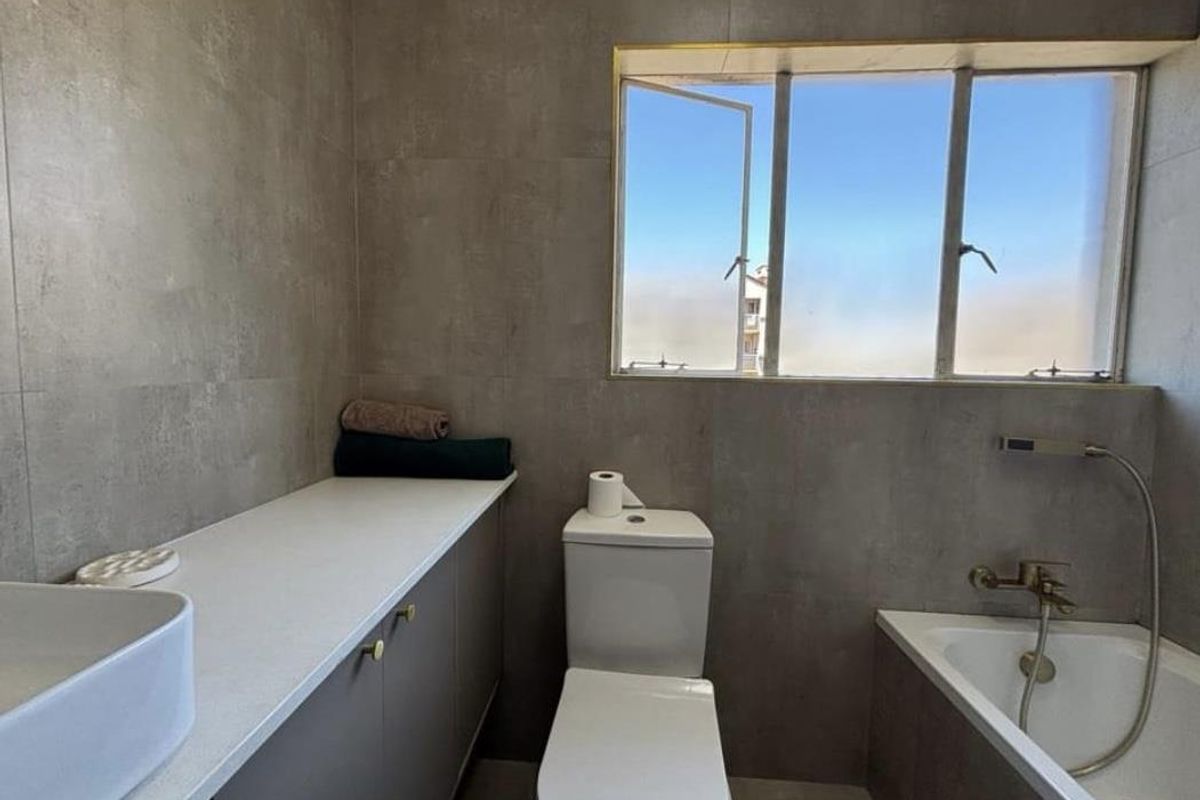 A modern bathroom featuring grey wall tiles, a built-in vanity with a countertop basin, a toilet, and a bathtub with a handheld shower fixture.