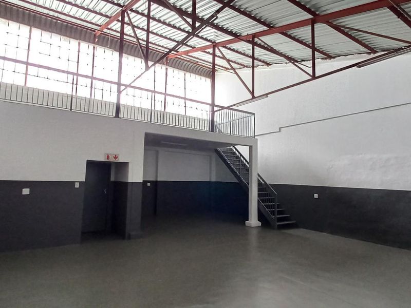 Spacious industrial unit featuring a mezzanine level, steel staircase, high ceilings with exposed trusses, and large windows providing ample natural light.