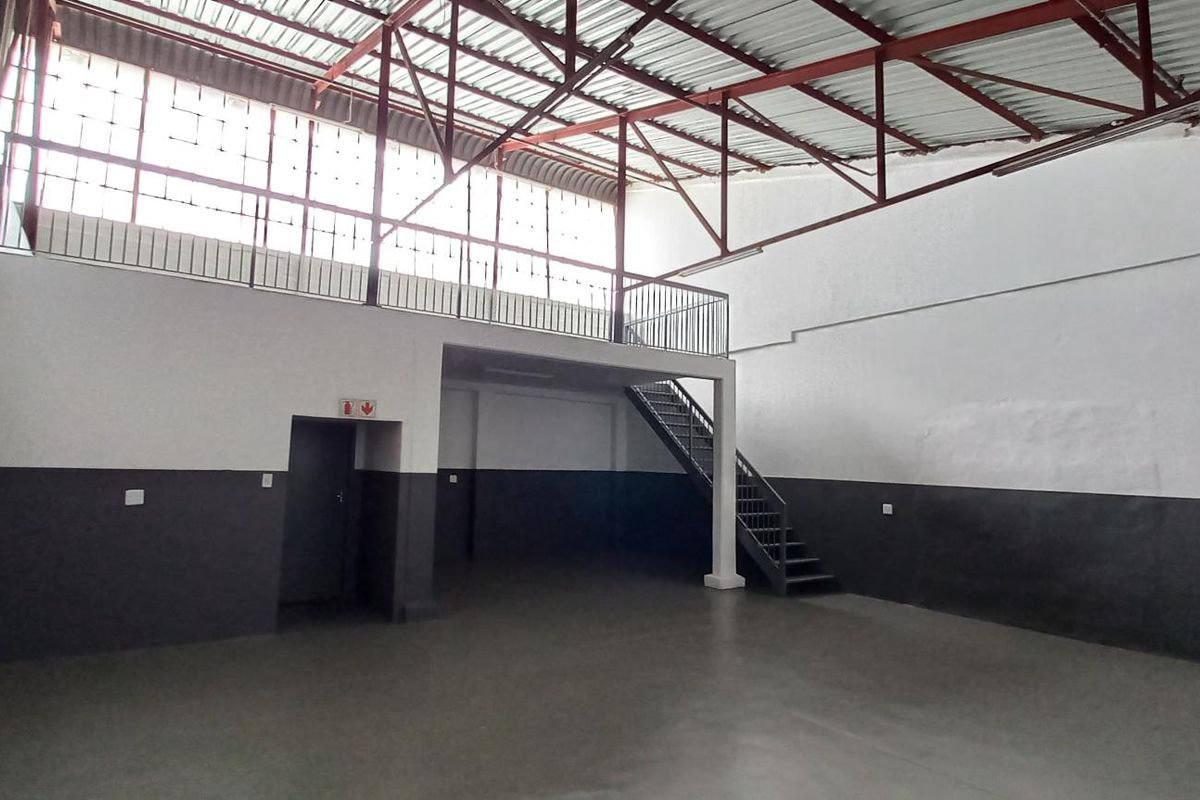 Spacious industrial unit featuring a mezzanine level, steel staircase, high ceilings with exposed trusses, and large windows providing ample natural light.