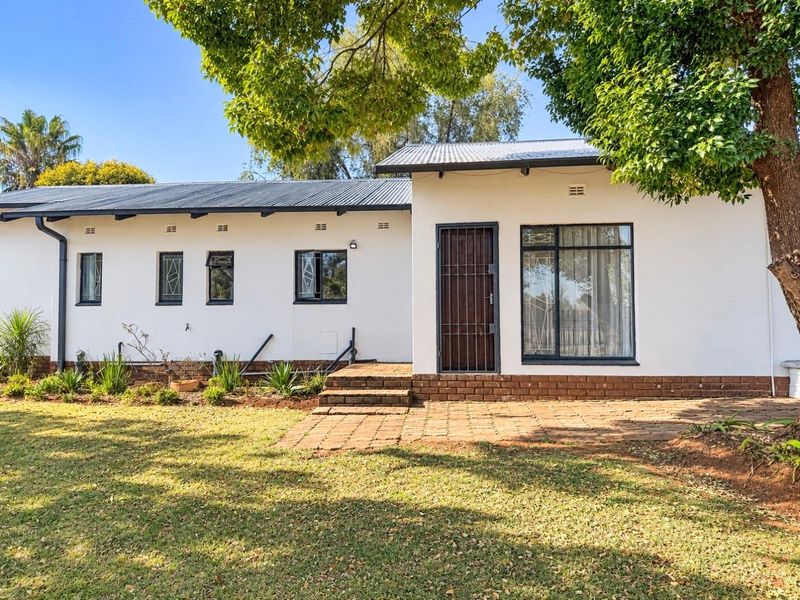Rear view of the house featuring a white facade, corrugated metal roof, brick paved patio area, and established garden surroundings.