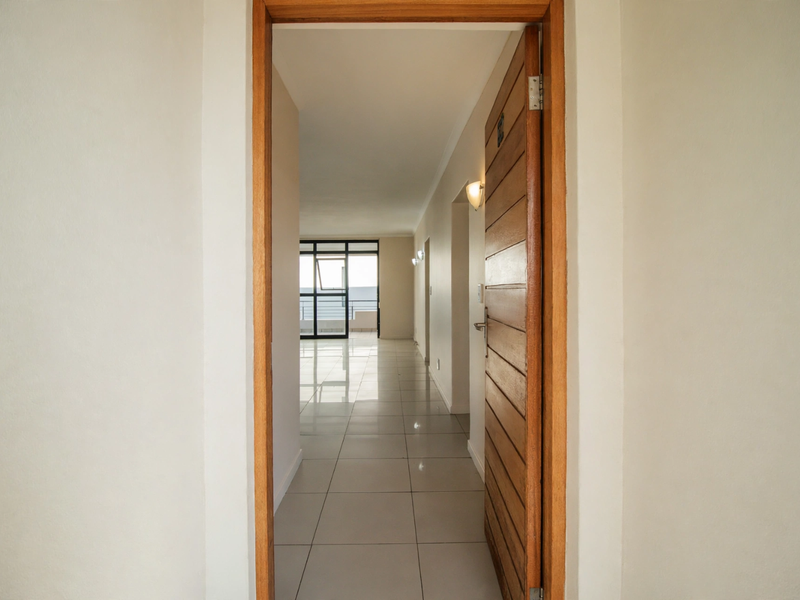 A bright, clean hallway with neutral tiled flooring and a wooden door, leading towards an open-plan living area with natural light.